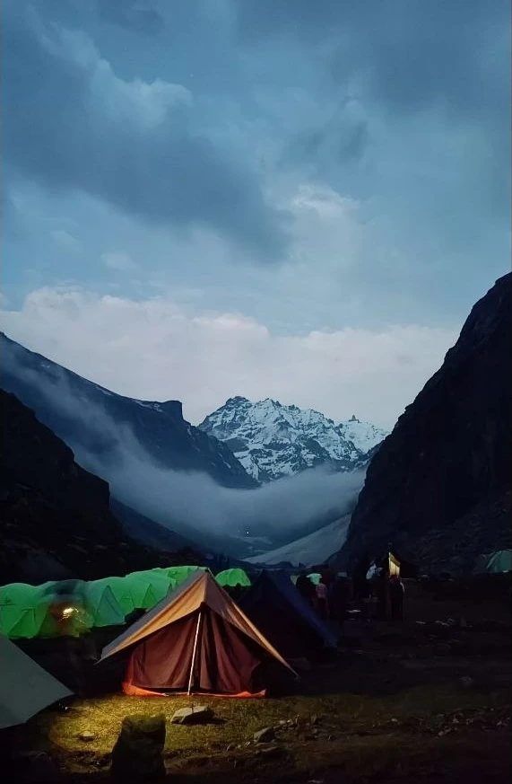 Tents lit up at a mountain camp at dusk, below a snow peak and low clouds